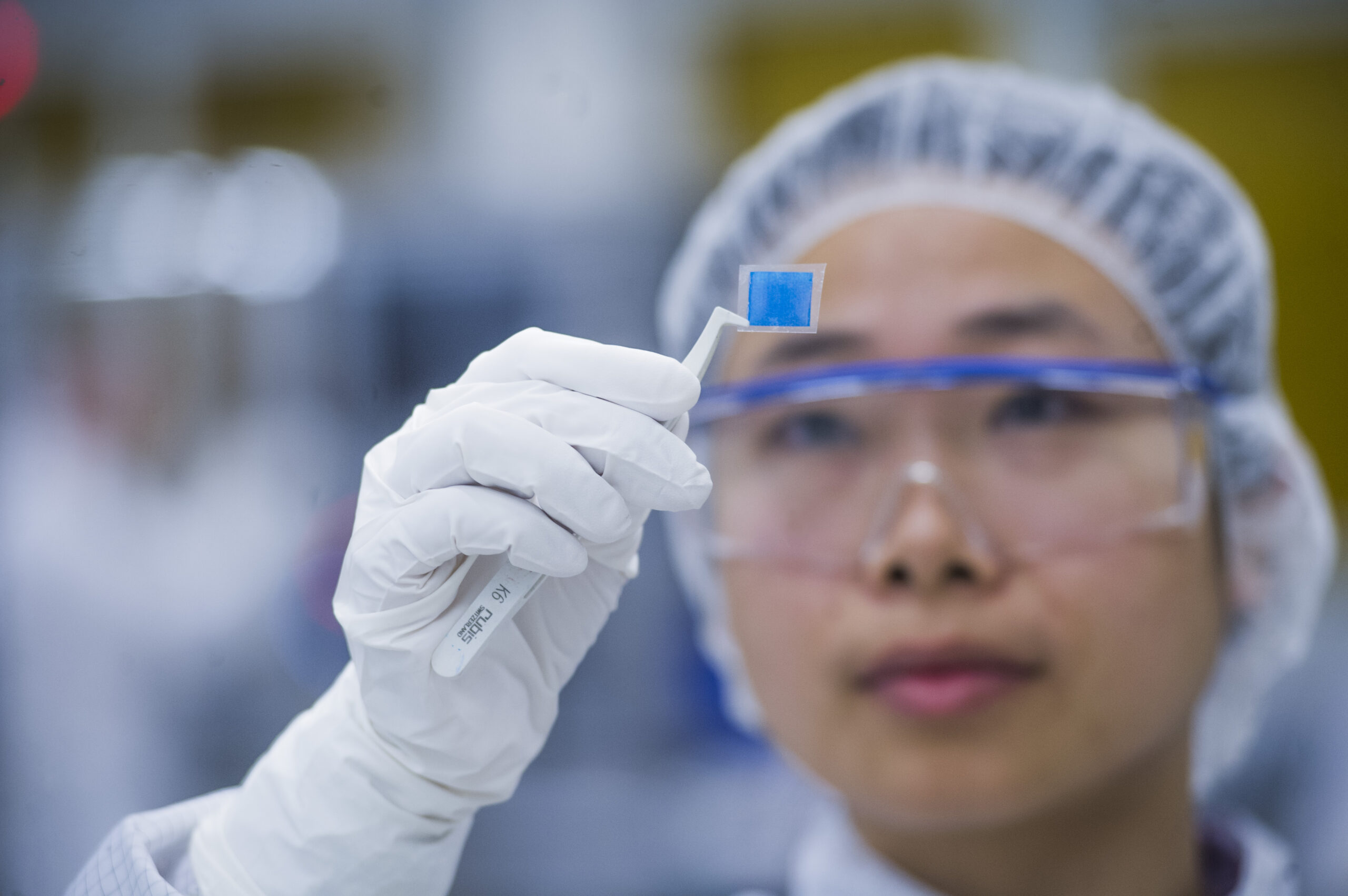 woman in cleanroom clothes and with safety glases checks a small piece of printed electronics she helds with a tweezer